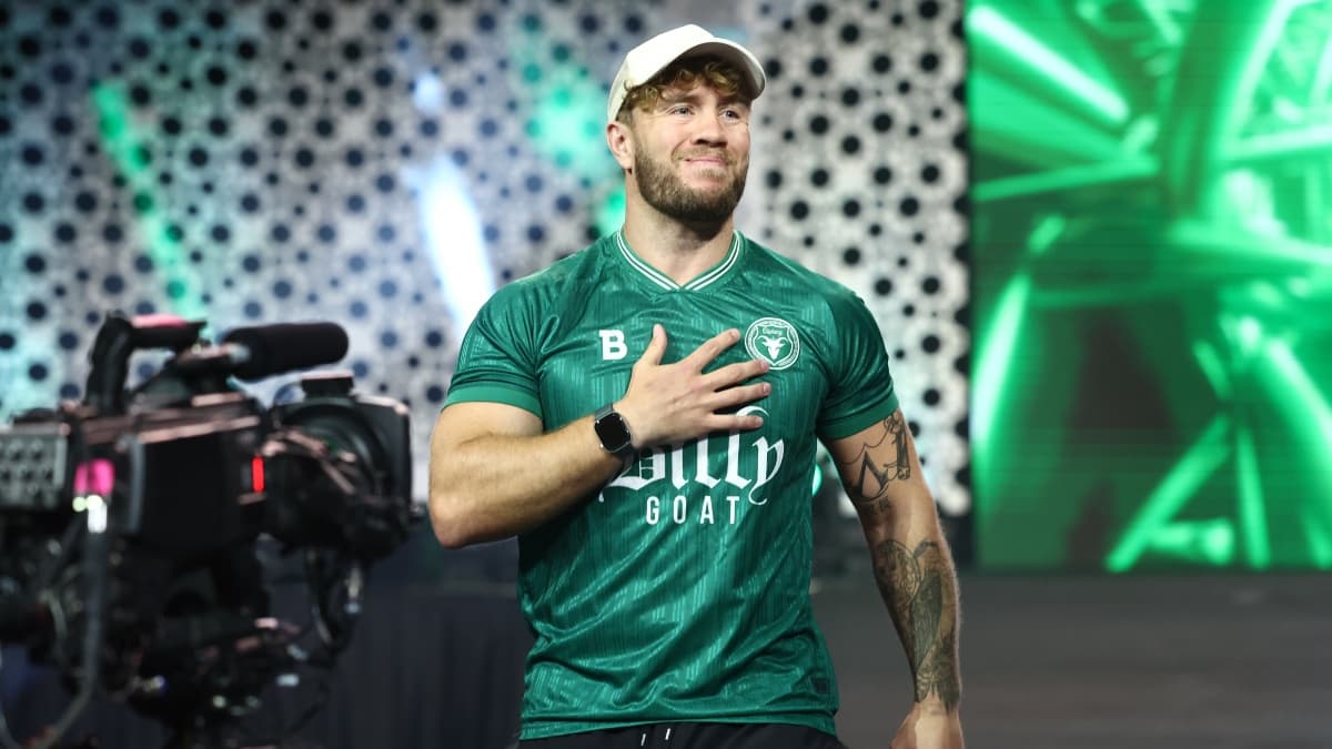 Will Ospreay wearing a green football shirt and white hat with his hand on his chest